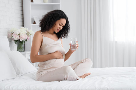 Young pregnant African-American woman with glass of water sitting in bedroomの写真素材