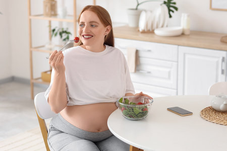 Young pregnant woman eating vegetable salad at table in kitchen