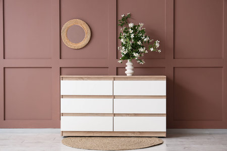 Vase with blooming jasmine flowers on chest of drawers near brown wall in interior of living room