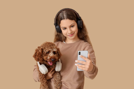 Teenage girl in headphones with Poodle dog using mobile phone on beige background