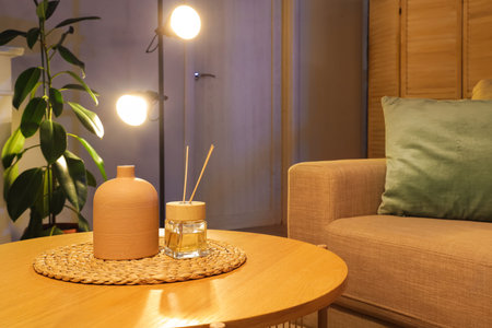 Wooden coffee table with vase, aroma sticks and wicker mat in stylish living room at evening