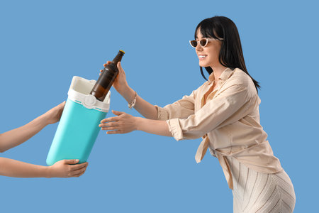 Young woman taking bottle of cold beer from portable refrigerator on blue backgroundの写真素材