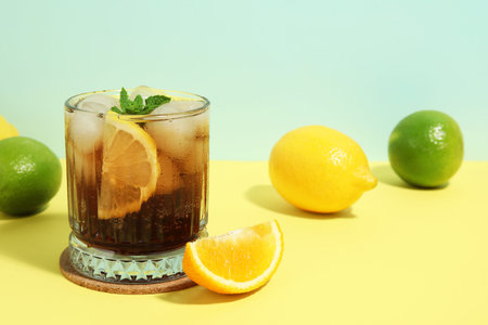 Glass of Long Island iced tea cocktail with mint and different citrus fruits on yellow table against...