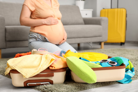 Young pregnant woman packing suitcase with clothes and travel accessories at homeの写真素材