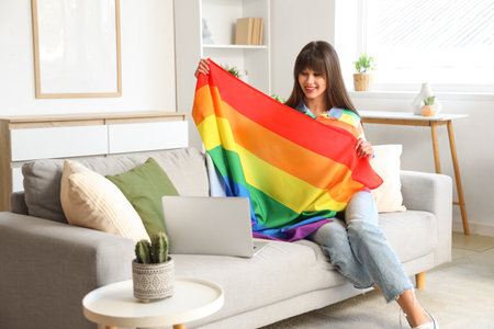 Beautiful young woman with LGBT flag and modern laptop sitting on sofa in living roomの写真素材