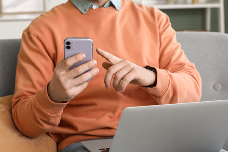 Young man with laptop using mobile phone on sofa at home, closeup