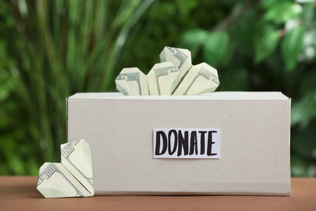 Box with the word DONATE and dollar banknotes in shape of heart on table outdoors
