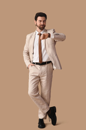 Handsome businessman in stylish suit looking at wristwatch on beige backgroundの写真素材