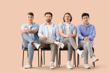 Group of young men sitting on chairs against beige background