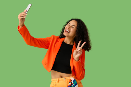 Stylish young African-American woman with mobile phone taking selfie on green backgroundの写真素材