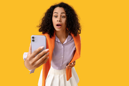 Shocked young African-American woman using mobile phone on yellow background