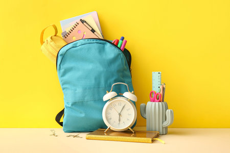 School backpack with alarm clock and stationery on yellow background
