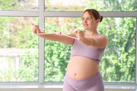 Sporty pregnant woman training with expander near window at gym