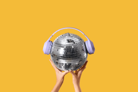 Female hands with disco ball and headphones on yellow background