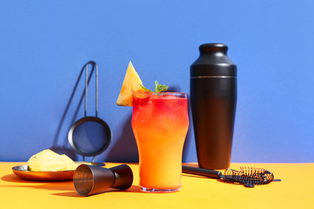 Glass of tasty cocktail garnished with pineapple and mint on orange table against blue background