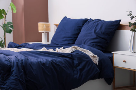 Bed with stylish blue bedding, bedside tables, lamp and houseplants in bedroom