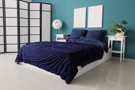 Bed with stylish blue bedding, bedside tables, folding screen and hanging paintings on wall in...