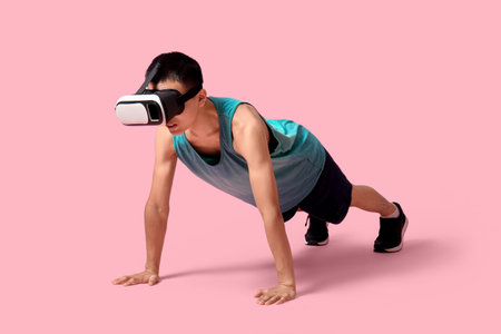 Sporty young Asian man in VR glasses doing push-ups on pink background