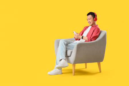 Young Asian man with headphones using mobile phone in gray armchair on yellow backgroundの写真素材