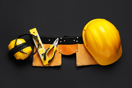 Hardhat, protective earmuffs, tool belt and construction tools on black background, top view