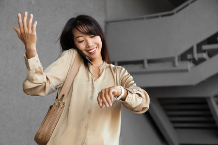 Happy Asian businesswoman with wristwatch talking by mobile phone outdoors. Time management concept