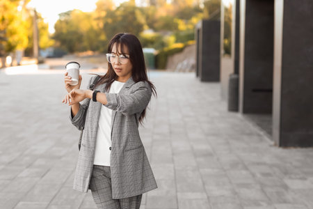 Young Asian businesswoman with coffee looking at wristwatch outdoors. Time management concept