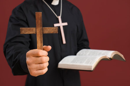 Young priest with wooden cross and Bible on red background, closeup