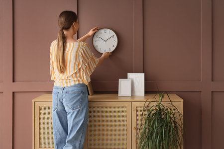 Young woman hanging clock on brown wall in room, back view