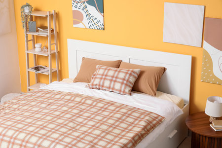 Comfortable bed, table and shelving unit near yellow wall in interior of bedroom, closeup