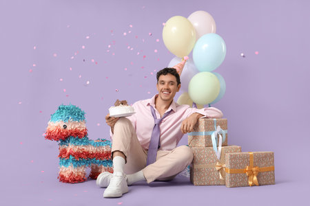 Happy young man with cake and gifts celebrating Birthday on lilac backgroundの写真素材