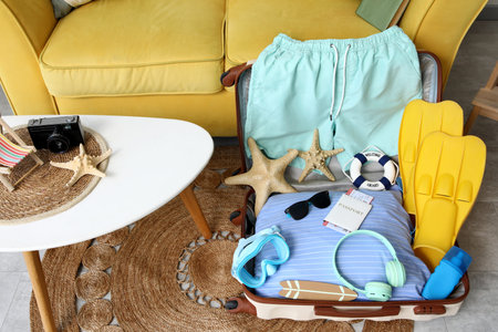 Open suitcase with men's swimwear, headphones and beach accessories on floor near sofa, closeup