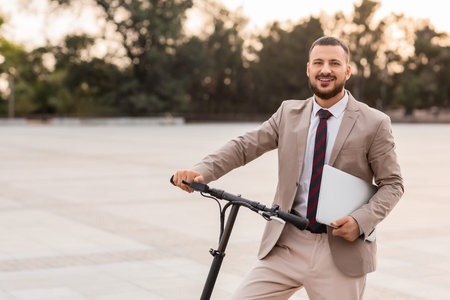 Handsome young businessman with laptop and electric scooter at city street