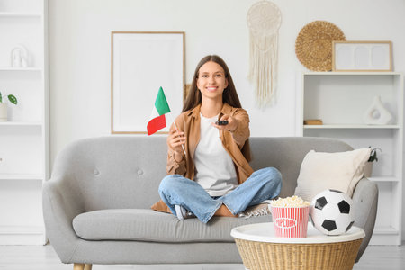Young woman with TV remote controller and Italian flag watching football on sofa at homeの写真素材
