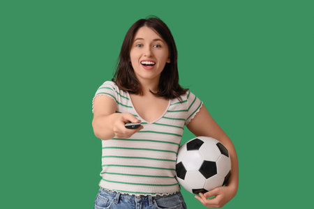 Young woman with TV remote controller watching football on green background