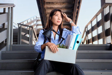Fired Asian businesswoman with personal stuff sitting on stairs outdoorsの写真素材