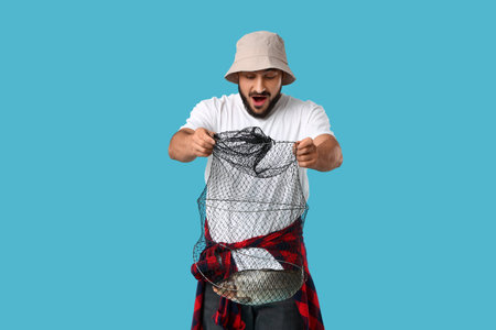 Surprised young man with caught fish in keepnet on blue backgroundの写真素材