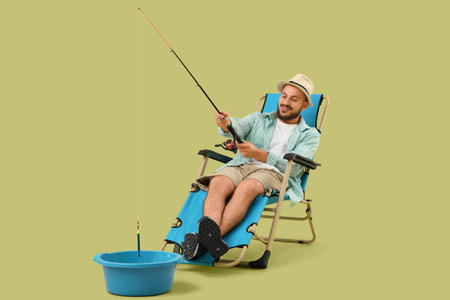 Young man sitting on folding chair and fishing in plastic basin against green backgroundの写真素材