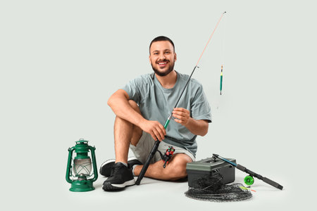 Handsome young man with fishing rod, tackle box and lantern sitting on gray backgroundの写真素材