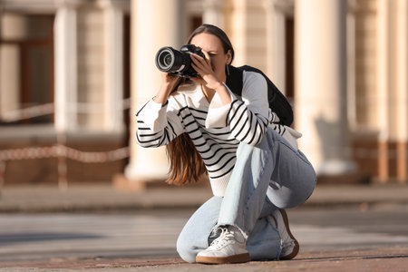 Young happy female photographer with camera and backpack sitting and taking photo on city street