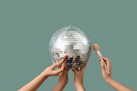 Female hands with disco ball, makeup brush and lipstick on green background