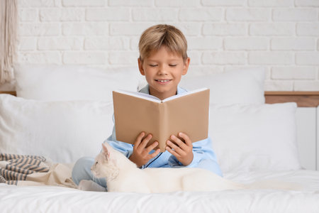 Cute little boy with cat reading book in bedroomの写真素材