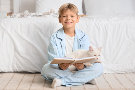 Cute little boy with cat reading book on carpet in bedroomの写真素材