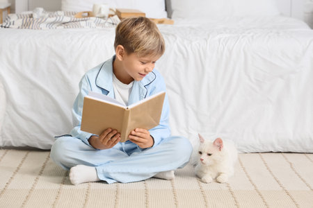 Cute little boy with cat reading book on carpet in bedroomの写真素材