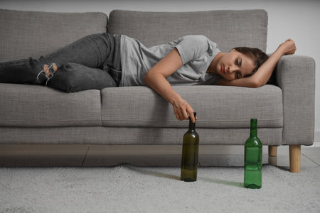 Sad young woman with bottles of alcohol lying on sofa at homeの写真素材