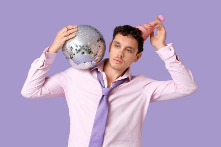 Young man in party hat with disco ball celebrating Birthday on lilac backgroundの写真素材