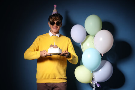 Happy young man with Birthday cake and balloons on dark blue backgroundの写真素材