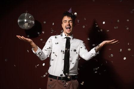 Happy young man with confetti and disco ball celebrating Birthday on burgundy backgroundの写真素材