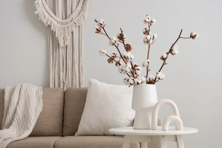 Vase with cotton flowers and statuette on table near white wall in living room, closeup