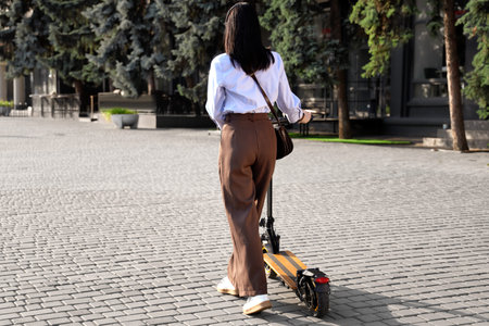 Young businesswoman in headset riding electric scooter outdoors, back view