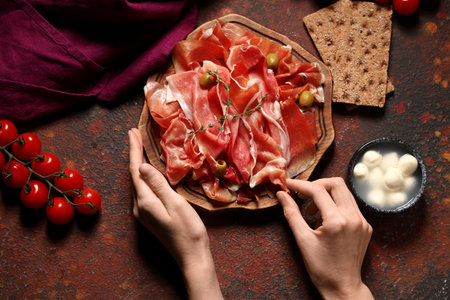 Female hands with wooden board of delicious prosciutto slices and mozzarella cheese on dark backgroundの写真素材